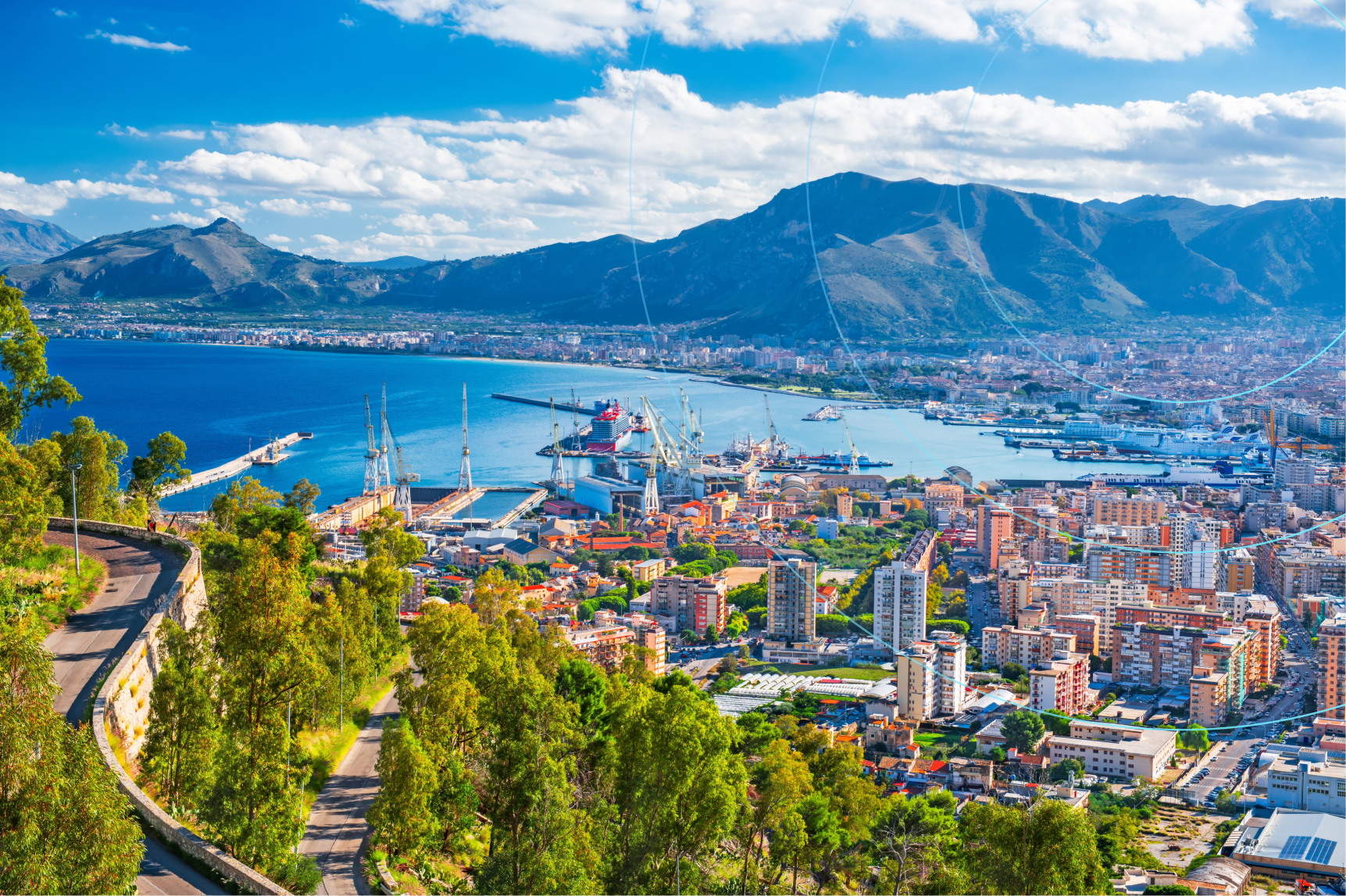 Panoramic view of a coastal city with a harbor, ships, and cranes, surrounded by mountains and overlooking the sea under a blue sky with clouds.