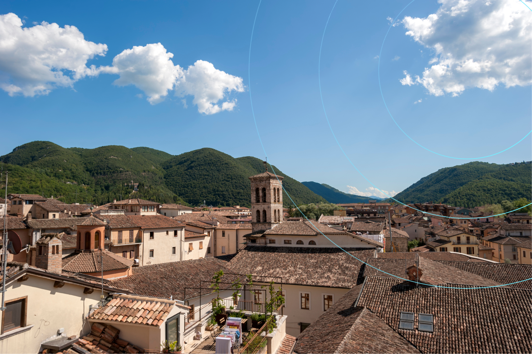 Panoramic view of an Italian village with tiled rooftops and a stone bell tower, surrounded by green hills under a blue sky with clouds.